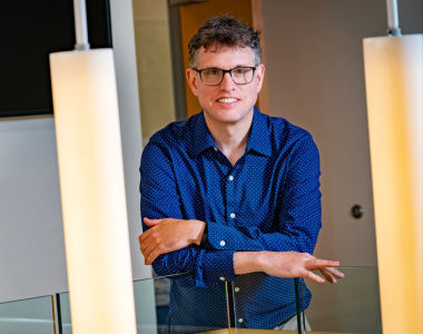 Man wearing glasses and a blue shirt poses behind hanging pendant lights